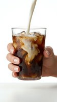 Pouring creamy milk into cola drink with ice cubes in glass held by hand against white background, suitable for beverage advertising and food photography.