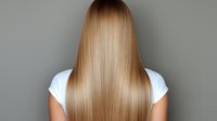 Young woman with long straight honey blonde hair shown from behind wearing white t-shirt against gray background, demonstrating healthy, shiny hair texture.