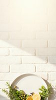 Cream brick wall background with empty round plate, fresh herbs, capers, and lemon slices for food styling and culinary design projects.