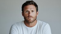 Confident Caucasian man with beard wearing white t-shirt looking at camera against light background. Professional headshot style portrait showing genuine expression.