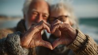 Senior couple making heart shape with hands together at beach, elderly man and woman showing love and romance in golden hour light by ocean.