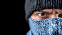 Intense close-up of Caucasian male eyes wearing winter knit beanie and face mask against black background, dramatic lighting emphasizes mysterious gaze and facial expression.