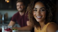 Young African American woman with curly hair smiling brightly at dinner table, wearing yellow top, with male companion and fresh berries in foreground, casual dining atmosphere.
