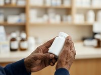 Senior male with dark skin tone holding white supplement bottle in pharmacy setting with shelves of products in background.