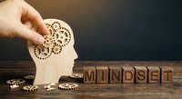 Hand placing wooden gear into human head silhouette with scattered cogs and MINDSET letter blocks on rustic table representing mental development.