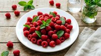 Fresh raspberries arranged on white plate with mint leaves, scattered berries and water glass on wooden table. Food styling for culinary, recipe and healthy eating content.
