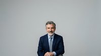 Middle-aged man with gray hair and facial hair, Caucasian ethnicity, wearing dark navy business suit and light blue dress shirt. Professional corporate portrait against neutral gray background.