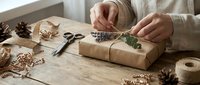 Woman wrapping eco-friendly gift with brown kraft paper and natural twine on rustic wooden table with pine cones and dried flowers for sustainable holidays.