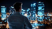 Young businessman in suit looking at illuminated night city skyline with modern skyscrapers and office buildings. Back view silhouette against urban lights.