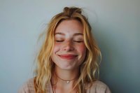 Young blonde woman with freckles and closed eyes smiling peacefully against light blue wall background, expressing joy and contentment in natural lighting.