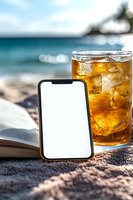 Smartphone mockup with blank screen next to iced tea glass and open book on beach. Tropical ocean background with bokeh lights creates relaxing summer atmosphere.