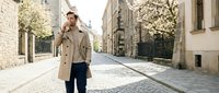 Young businessman in beige coat talking on phone while walking down cobblestone street in historic city center during spring season.