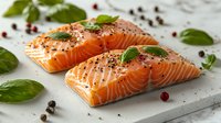 Raw salmon fillets seasoned with black pepper and fresh basil leaves on white marble board, garnished with pink peppercorns and herbs for cooking preparation.