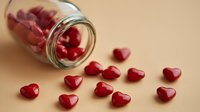 Red heart-shaped candies spilling from glass jar onto beige background, creating romantic composition for Valentine's Day celebrations or love-themed designs.