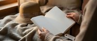 Woman reading open book while relaxing on cozy sofa with warm blanket and straw hat nearby creating peaceful home atmosphere for leisure time.