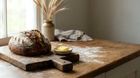 Freshly baked sourdough loaf with scoring marks on wooden cutting board, artisan bread with butter and dried wheat arrangement for bakery and food styling.