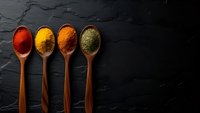 Four wooden spoons filled with colorful ground spices including paprika, turmeric, and herbs arranged on dark slate background for culinary cooking.