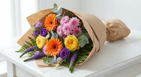 Colorful mixed flower bouquet with orange gerberas, purple roses, yellow ranunculus and pink carnations wrapped in kraft paper on white surface.