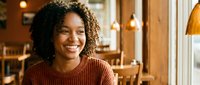 Young African American woman with curly hair smiling warmly in cozy cafe restaurant interior with wooden decor and warm lighting for lifestyle content.