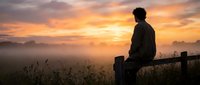 Young man sitting on a fence, gazing at a dramatic misty sunrise over a rural field, warm golden sky, peaceful solitude.