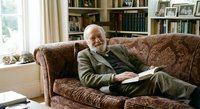 Senior Caucasian man with white beard relaxing on vintage leather sofa in cozy home library with bookshelves and natural window light for lifestyle content.