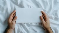 Hands holding blank white card or paper mockup against white crumpled bedsheet background, ready for custom text or design.