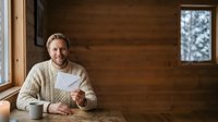 Middle-aged Caucasian man with beard wearing casual cardigan sitting at wooden table with coffee mug and notebook in rustic cabin interior, suitable for lifestyle and remote work concepts.