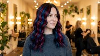 Woman with long wavy dark hair featuring blue and purple highlights wearing gray sweater in festive indoor venue with warm string lights and holiday decorations.