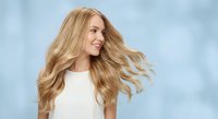 Young blonde woman with flowing wavy hair in motion against soft blue sky background, expressing joy and freedom in natural outdoor portrait.
