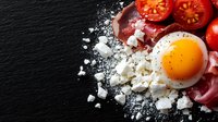 Fresh sunny side up egg with crumbled feta cheese, sliced red tomatoes, and sea salt scattered on dark slate background, top view food photography.