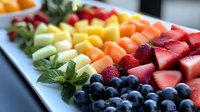 Fresh colorful fruit platter with strawberries, blueberries, melon, pineapple and mint garnish arranged in diagonal rows on white serving plate for catering.