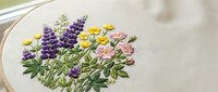 Delicate wildflower embroidery on natural linen fabric featuring purple lupines, yellow daisies and pink blooms in wooden hoop for handcraft projects.