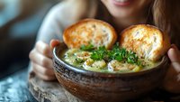 Woman holding rustic bowl of creamy soup with fresh herbs and crusty bread rolls for cozy dining experience.
