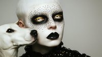 Avant-garde portrait of pale woman with dotted face paint and black lips posing with white dog against neutral background for artistic fashion concepts.