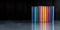 Colorful row of hardcover books standing on reflective dark surface against black background, creating minimalist composition with rainbow spectrum effect.
