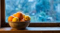 Rustic wooden bowl with fresh oranges and lemons on a windowsill against a rain-covered window, cozy autumn mood.