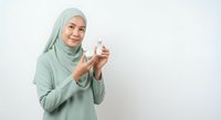Young Muslim woman in green hijab holding glass of milk smiling at camera on white background for healthy lifestyle and nutrition concepts.