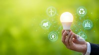 Businessman hand holding glowing LED bulb with green energy icons including solar, wind, and recycling symbols on nature background.