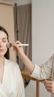 Professional makeup artist applying powder blush to young Caucasian woman's cheek with soft brush in bright studio setting.