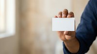Businessman in dark blue shirt holding blank white business card with hand, isolated on blurred office background for corporate branding and contact information display.