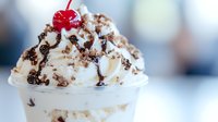 Creamy vanilla milkshake topped with whipped cream, chocolate sprinkles and maraschino cherry in clear glass, close up view on light background. Food photography.