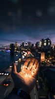 Hand holding glowing heart-shaped light against blurred city skyline at dusk. Warm bokeh lights reflect on water with illuminated skyscrapers in background.