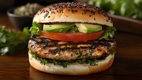 Grilled chicken burger with fresh vegetables and black sesame seeds on brioche bun, layered with lettuce, tomato and cucumber slices on wooden surface.