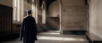 Graduate in black cap and gown walking through historic university corridor with arched windows and stone architecture for academic achievement concepts.