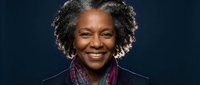 Professional African American businesswoman with curly gray hair smiling confidently in dark suit against navy background for corporate presentations.