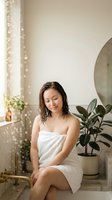 Young Asian woman relaxing in modern bathroom wrapped in white towel with wet hair enjoying peaceful spa moment surrounded by plants.