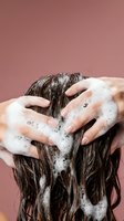 Woman washing long brown hair with shampoo foam creating bubbles during hair care routine against pink background for beauty and hygiene concepts.