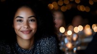 Young African American woman with natural curly hair smiling warmly at camera in evening setting with soft bokeh lights and festive atmosphere in background.