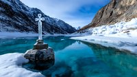 Cross monument on rock overlooking turquoise mountain lake surrounded by snow-covered peaks and cliffs, dramatic winter landscape with blue sky reflections in water.