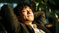 Young Asian man relaxing with hands behind head, eyes closed in peaceful moment. Sunlight streaming through window creates warm atmosphere for wellness content.
